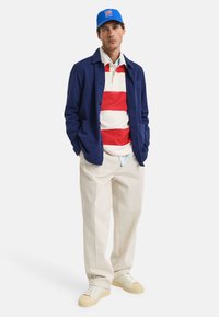 Marineblauwe jacket over een rood-wit gestreepte polo, beige broek en witte sneakers met rubberen zolen; blauwe pet met logodetail.