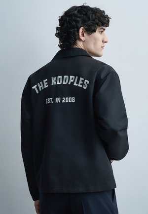 Άνδρας με σγουρά μαλλιά που φοράει μαύρο μπουφάν με την κεντητή επιγραφή "THE KOOPLES EST. IN 2008" στην πλάτη, στραμμένος πλάγια.