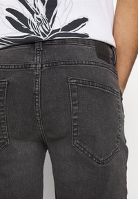 Persona vistiendo jeans de mezclilla negra desgastada y una camisa blanca con un gran estampado floral negro visible en la cintura.