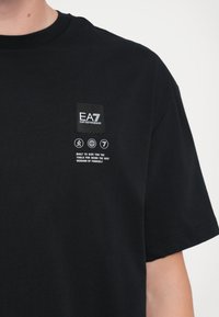 Melna kokvilnas T-krekls ar nelielu taisnstūrveida logotipa plāksteri, ikonām un tekstu. Īsas piedurknes ar brīvu piegriezumu. Vienkāršs dizains, bez rakstiem.