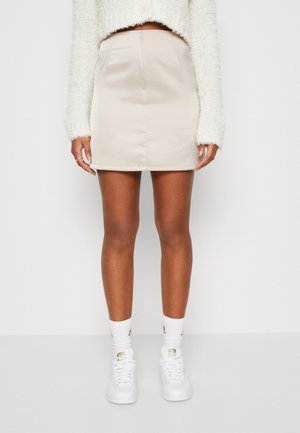 Kvinde iført beige mininederdel, hvidt blødt strik, hvide sokker og hvide sneakers mod en ensfarvet hvid baggrund.