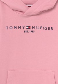 Růžová bavlněná mikina s kapucí a kangaroo kapsou, s logem "TOMMY HILFIGER EST. 1985" v tmavě modré barvě a malou červenobílou emblém.