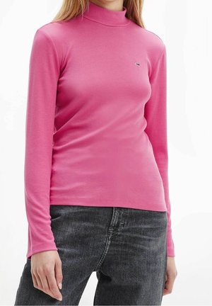 Femme portant un haut moulant à manches longues rose côtelé avec un col montant et un jean noir, debout devant un fond blanc.