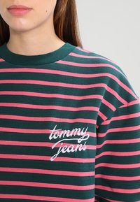 Sudadera a rayas verdes y rosas con cuello redondo, con la inscripción "tommy jeans" bordada en blanco en el frente. Textura suave de algodón.