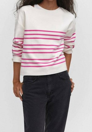 Personne portant un pull blanc à rayures horizontales roses et un jean taille haute foncé, les mains dans les poches, devant un fond uni.