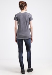 T-shirt grigio a maniche corte con orlo leggermente curvo, abbinato a jeans skinny scuri. Il modello indossa scarpe con zeppa nere e volge le spalle alla fotocamera.