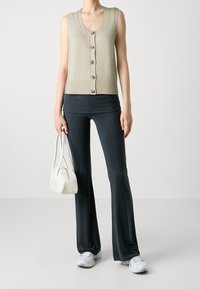 Chaleco de punto color beige claro con dobladillo acanalado y seis botones en la parte delantera, combinado con pantalones de campana oscuros y ajustables, y un pequeño bolso blanco. Zapatillas blancas.