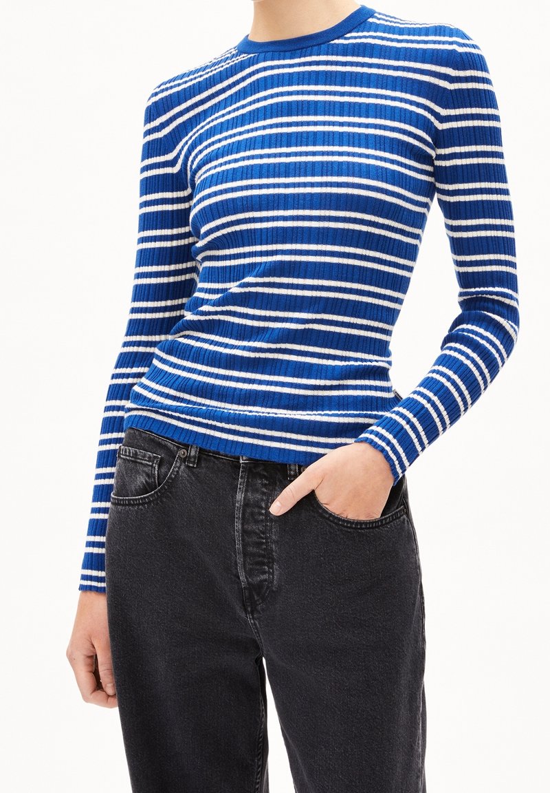 Personne portant une chemise rayée bleu et blanc à manches longues rentrée dans un jean noir taille haute, une main dans une poche.