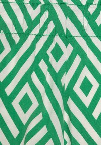Tissu vert et blanc présentant un motif géométrique de diamants et de lignes en zigzag. Le matériau semble léger avec une texture lisse.