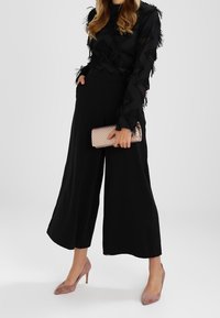 Svart långärmad blus med fransdetaljer, tillsammans med vida svarta byxor. Håller en ljusrosa randig clutch och bär rosa klackar.