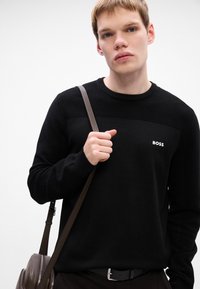 Mladý muž v čiernom svetri BOSS, ktorý drží popruh hnedého koženého batohu na svetlom pozadí.