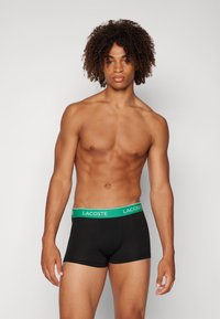 Black boxer briefs with a green waistband featuring the "LACOSTE" logo. The fabric appears smooth and stretchy with a fitted design.
