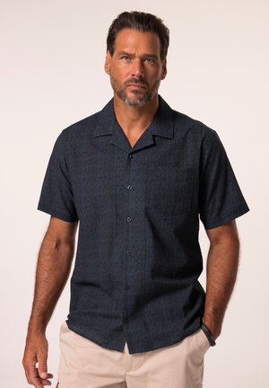 Man met kort donker haar en baard, dragend een donker patroon korte mouwen button-up overhemd en licht beige broek tegen een effen achtergrond.