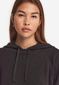 Schwarzer Hoodie aus weichem Stoff, mit Kordelzug, gerippten Bündchen und relaxter Passform. Nahaufnahme des Gesichts des Modells, neutraler Hintergrund.