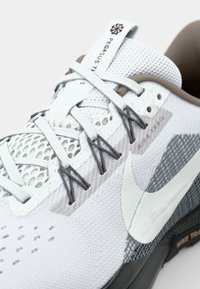 Gros plan sur une chaussure de course Nike Pegasus Trail blanche avec empeigne en mesh, câbles Flywire gris et logo Nike swoosh sur le côté.