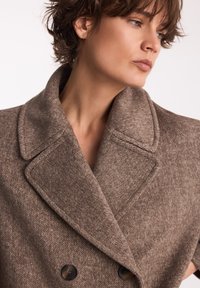 Dwurzędowy płaszcz wełniany w brązowy wzór w jodełkę. Posiada szerokie klapy i dwa czarne guziki z przodu. Miękka tekstura i wyraźny kształt.