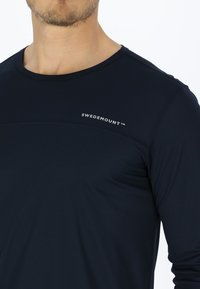Man draagt een marineblauw shirt met lange mouwen met het "Swedemount"-logo op de bovenborst tegen een witte achtergrond.