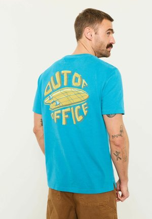 Mann mit Tattoos, der ein blaues Hemd mit gelbem "OUT OF OFFICE"-Text und einer Skateboard-Grafik auf dem Rücken trägt, steht vor einem weißen Hintergrund.