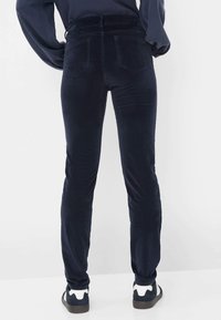 Pantalon en velours côtelé bleu marine avec une coupe slim et des poches à l'arrière, présentant une texture douce et une taille haute. Associé à des chaussures foncées.