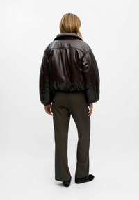 Eine Frau mit blonden Haaren trägt eine dunkelbraune glänzende Daunenjacke, graue Hosen und schwarze Schuhe und steht mit dem Rücken zu einer weißen Hintergrund.