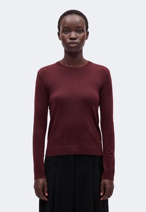 Jeune femme aux cheveux courts portant un pull bordeaux ajusté à manches longues et une jupe noire sur un fond clair uni.