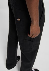 Pantalones cargo negros de lona con múltiples bolsillos, textura duradera y un parche con el logo de Dickies en el bolsillo trasero. Diseño minimalista.