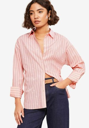 Camicia a maniche lunghe di colore rosa con strisce verticali bianche e piccoli dettagli. Presenta un colletto e maniche arrotolate. Abbinata a jeans scuri.