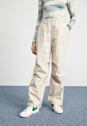 Pantalon large beige en coton, avec une taille à cordon, deux poches latérales et une texture subtile. Associé à des baskets blanches.