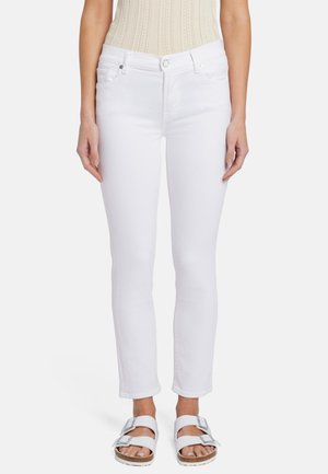 Femme portant un jean slim blanc, un haut sans manches beige en maille et des sandales blanches à double bride, debout devant un fond uni.