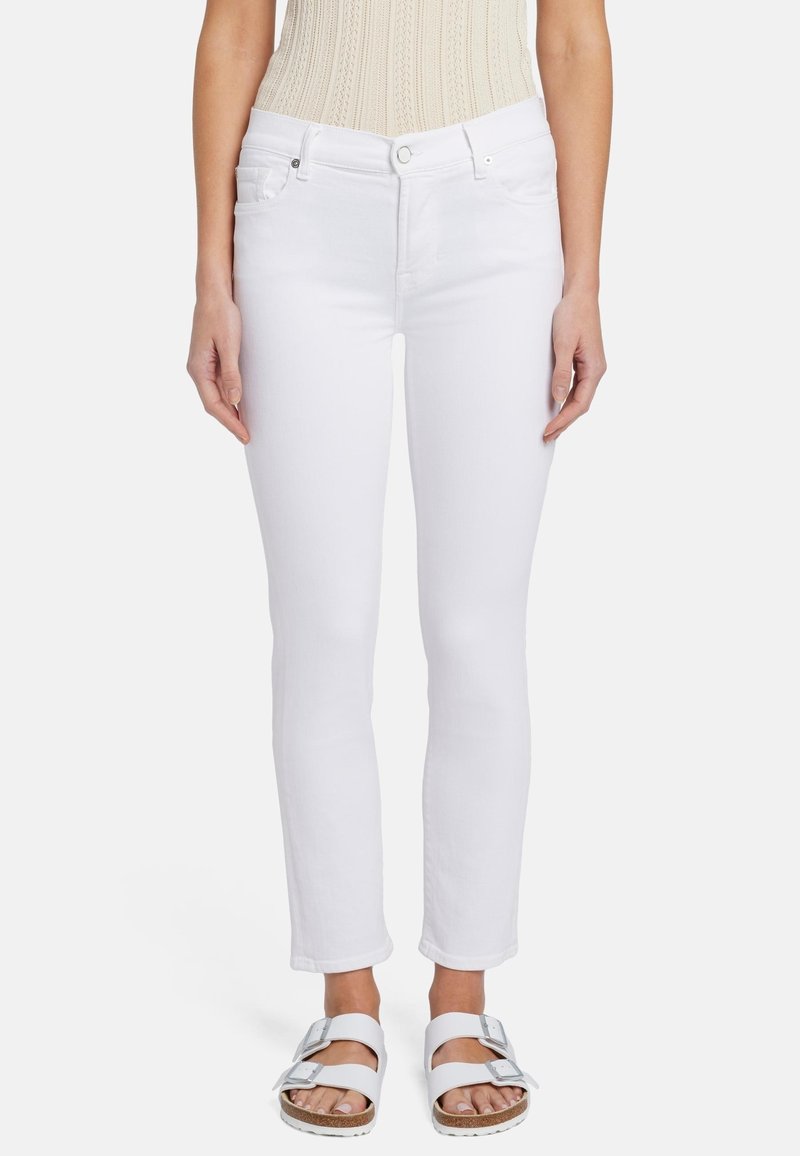 Femme portant un jean slim blanc, un haut sans manches beige en maille et des sandales blanches à double bride, debout devant un fond uni.
