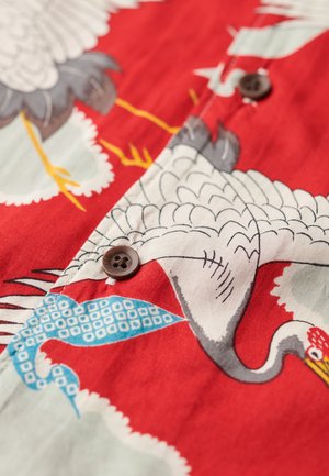 Tissu rouge avec un motif de grue, présentant des accents bleus et gris. Des boutons marron sont visibles, avec une texture lisse et des illustrations détaillées.