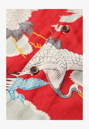 Tissu rouge avec un motif de grue, présentant des accents bleus et gris. Des boutons marron sont visibles, avec une texture lisse et des illustrations détaillées.