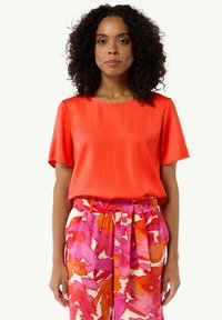 Jasnoczerwona bluzka z krótkim rękawem z gładkiego materiału, zestawiona z różowymi spodniami w wzory z wiązanym paskiem w talii i abstrakcyjnym kwiatowym wzorem.