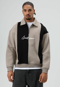 Lichtgrijze en zwarte sweatvest met rits en kraag, voorzien van een opvallend "Saut essence" logo aan de voorkant en geribbelde boorden.