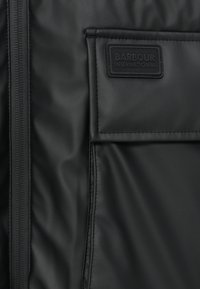 Εσωτερικό μαύρου μπουφάν από νάιλον με υφή, τσέπη με φερμουάρ και έντονη μαύρη ετικέτα με την επιγραφή "BARBOUR INTERNATIONAL".