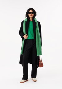 Sort frakke, grøn tørklæde, grøn top, sorte bukser og cremede sneakers. Modellen holder en brun håndtaske. Solbriller fuldender looket.