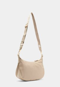 Beige Schultertasche mit einer geschwungenen Form und einem einzigen verstellbaren Trageriemen, ausgestattet mit einem Reißverschluss und goldenen Hardware-Akzenten.
