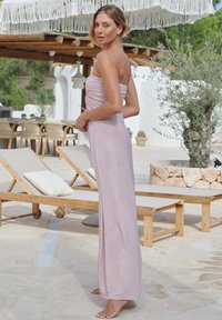 Trägerloses, bodenlanges Kleid aus einem hellrosa Stoff mit glatter Textur und gerafftem Design. Vor einem Innenhof mit Liegestühlen platziert.