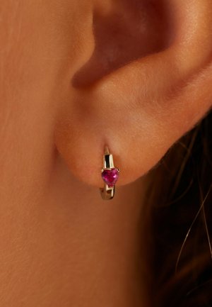 Gouden oorring in de vorm van een ring met een hartvormige roze edelsteen. Glad oppervlak, klein formaat en een veilig sluitmechanisme. Close-up van bij het oor.