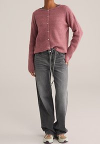 Růžový pletený svetr s knoflíky, dlouhými rukávy a volným střihem, doplněný volnými šedými džíny a vícero barevnými teniskami.