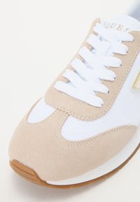 Baskets beiges et blanches avec une tige en daim, lacets blancs et une semelle en caoutchouc avec des accents tan. Détail du logo doré sur le côté.