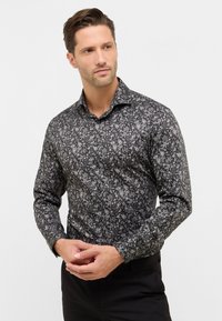Man in een zwart met bloemenpatroon langemouwshirt en zwarte broek, kijkt zijwaarts met de handen gekruist op de taille.