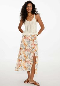 Krémový pletený top bez rukávů s krajkovým vzorem; dlouhá sukně s tiskem mušlí v oranžové, šedé a červené barvě; boční rozparek; hnědé sandály na ploché podrážce.