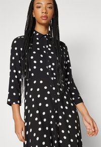 Robe noire à pois blancs, devant à boutons, col et manches trois-quarts. Tissu doux avec une coupe décontractée.