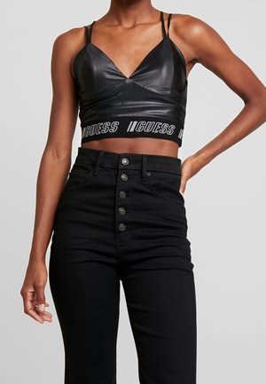 Vrouw draagt een zwarte cropped tanktop van nepleer met de tekstband "GUESS" en hooggetailleerde zwarte jeans met knopen, hand op heup.