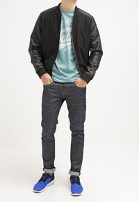 Černá bomber bunda s koženými rukávy, tyrkysové tričko s potiskem, tmavé džíny a modré sportovní boty s černými akcenty.
