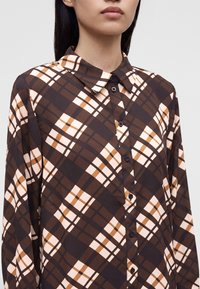 Femme portant une chemise à manches longues à motif écossais marron, beige et noir, visible du cou à la taille.