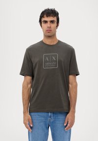 Camiseta de algodón verde oliva con un logo rectangular que dice "A|X ARMANI EXCHANGE" en el centro. Cuello redondo y mangas cortas.