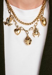 Zelta tonēta slāņota kaklarota ar vardes, sirds, ozola māsiņas, beetles un liesmas piespraudēm, ar teksturētu apdari uz baltas blūzes.