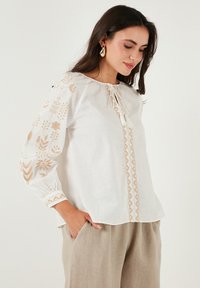 Blusa di lino bianca con ricami floreali beige e un fiocco decorativo sul davanti. Presenta maniche lunghe con polsini arricciati. Vestibilità casual e comoda.